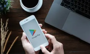 Ręce trzymają smartfon z logo Google Play, gotowe by dowiedzieć się, jak zaktualizować usługi Google Play. Obok laptop i kawa.