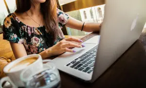 Kobieta w kwiecistej bluzce pracuje na laptopie, pijąc kawę. Uczy się, jak zapisać plik HTML w Notatniku.