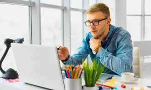 Młody mężczyzna w okularach uczy się, jak zrobić stronę HTML w notatniku, pracując przy laptopie.