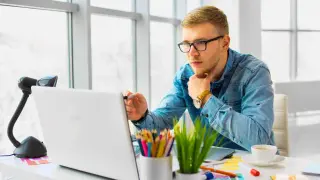 Młody mężczyzna w okularach uczy się, jak zrobić stronę HTML w notatniku, pracując przy laptopie.
