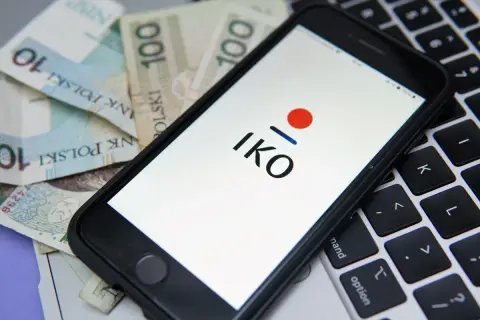 Chcesz wiedzieć, jak założyć IKO PKO? Na ekranie telefonu widzisz logo IKO, a obok polskie banknoty i klawiaturę laptopa.