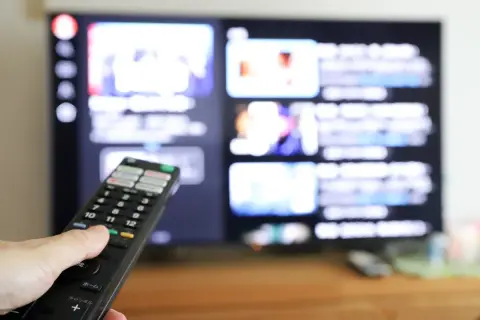 Dłoń z pilotem przed ekranem telewizora, na którym widać menu aplikacji. Dowiedz się, jak ściągnąć aplikację na telewizor.