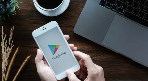 Ręce trzymają smartfon z logo Google Play, gotowe by dowiedzieć się, jak zaktualizować usługi Google Play. Obok laptop i kawa.