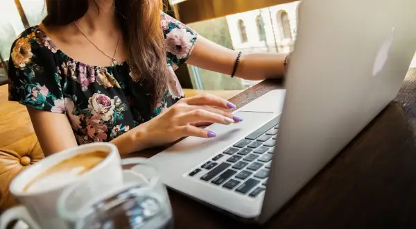 Kobieta w kwiecistej bluzce pracuje na laptopie, pijąc kawę. Uczy się, jak zapisać plik HTML w Notatniku.