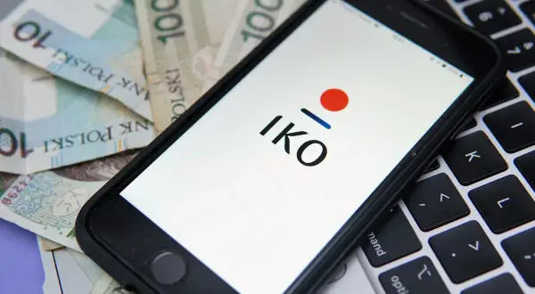 Chcesz wiedzieć, jak założyć IKO PKO? Na ekranie telefonu widzisz logo IKO, a obok polskie banknoty i klawiaturę laptopa.