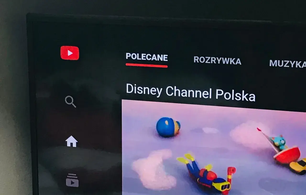 Ekran telewizora z aplikacją YouTube. Widoczne logo YouTube, menu "Polecane", "Rozrywka", "Muzyka" oraz kanał "Disney Channel Polska". Dowiedz się, jak zaktualizować aplikację YouTube.