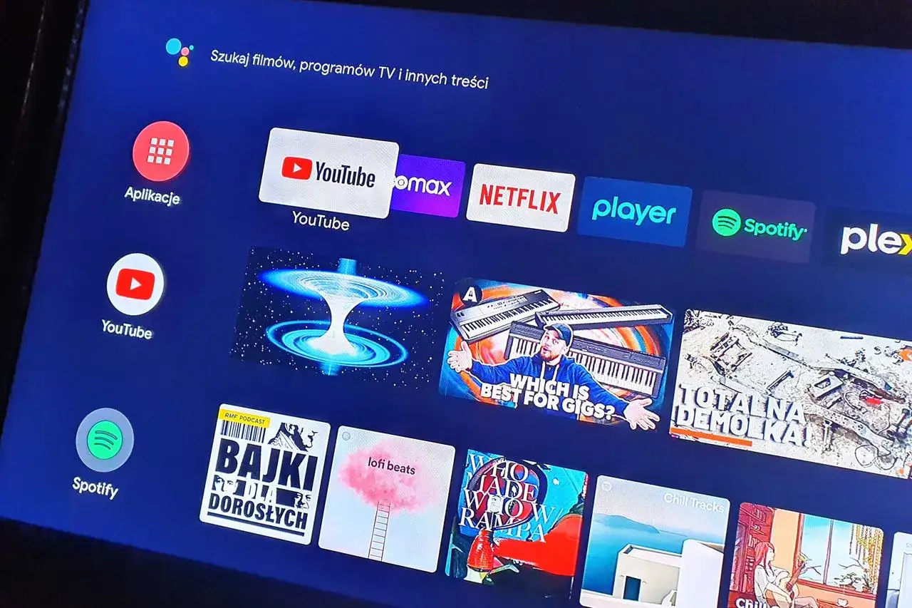 Ekran telewizora z ikonami aplikacji: YouTube, HBO Max, Netflix, Player, Spotify. Widoczny też przycisk 