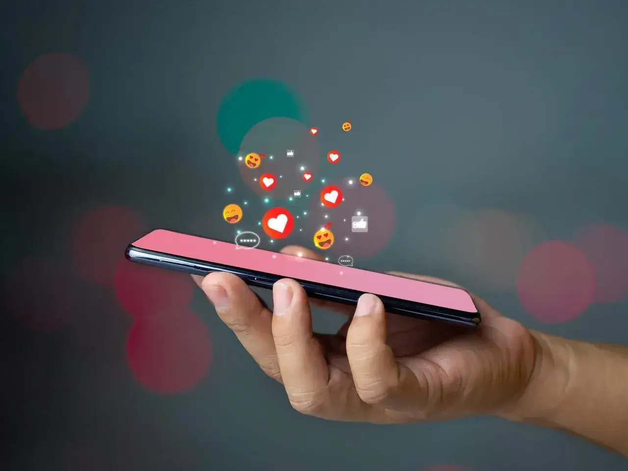 Dłoń trzyma iPhone'a, z którego unoszą się ikony polubień i emotikony. Dowiedz się, jak pobierać aplikacje na iPhone.