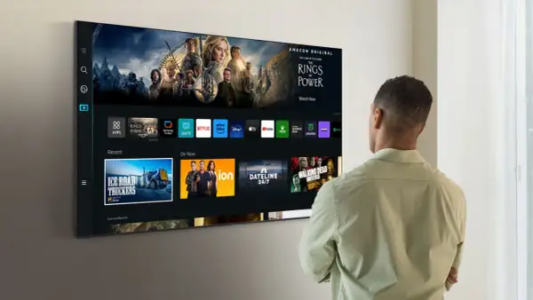 Mężczyzna patrzy na ekran telewizora Samsung Smart TV, gdzie wyświetlane są aplikacje i filmy. Dowiedz się, jak zaktualizować telewizor Samsung Smart TV, aby cieszyć się najnowszymi funkcjami.